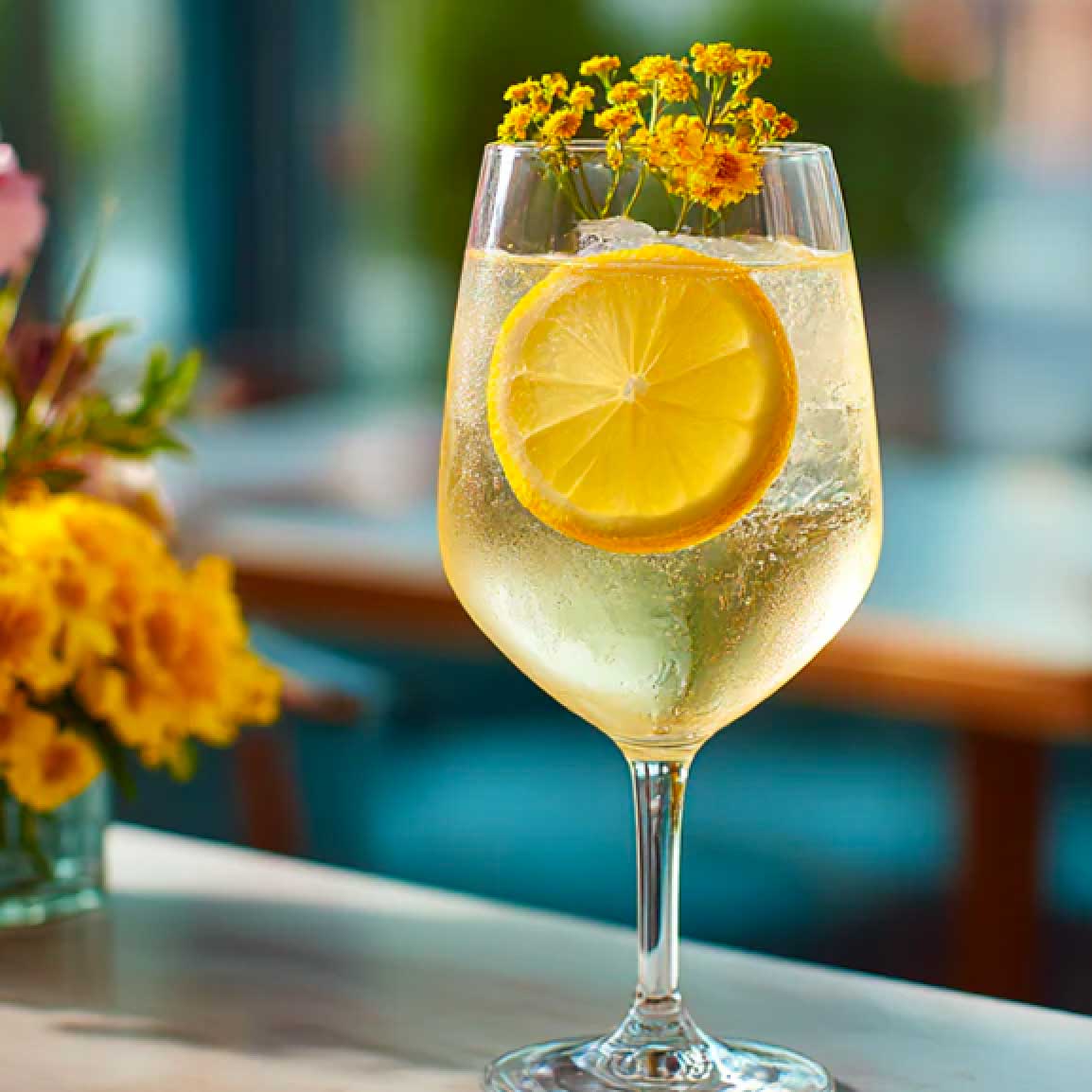 Cocktail Immortelle Vermouth & Tonic servi dans un verre de vin, cocktail aux couleur jaune, servi avec en décoration une tranche de citron jaune, de la glace et une tige de fleur jaune type mimosa. Ce cocktail est posé sur un comptoir en marbre de bar parisien. on voit en fonds une grande table rectangle de restaurant.